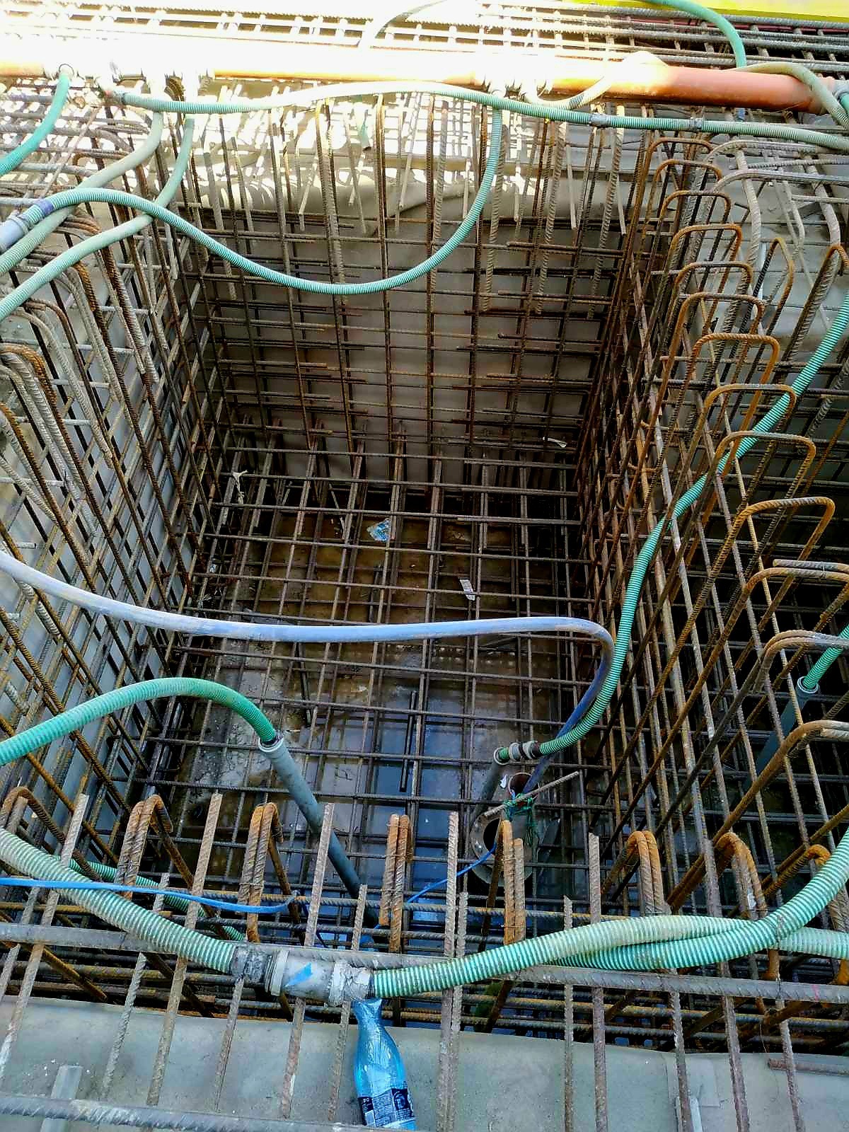 Głęboki wykop zbrojony stalowymi prętami, wypełniony wodą i poprzecinany zielonymi wężami, widok z góry.