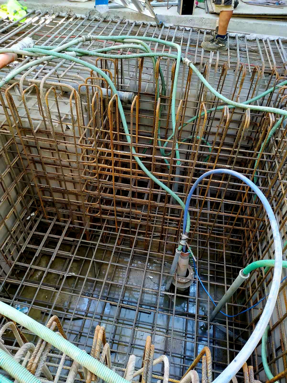 Siatka zbrojeniowa w wykopie fundamentowym, z zielonymi wężami do odwadniania i pompą zanurzeniową na dnie.