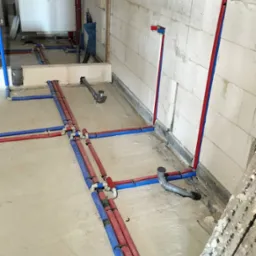 Rozprowadzenie instalacji wodnej i kanalizacyjnej w surowym wnętrzu. Czerwone i niebieskie rury prowadzone po podłodze i ścianach z betonu komórkowego.