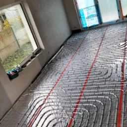 Białe rury ogrzewania podłogowego, zamocowane na siatce, ułożone w nowym pomieszczeniu z oknami. Widoczne narzędzia w tle. Czerwone pasy na podłożu.