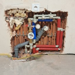 InstalHydroMax - Odsłonięta instalacja wodna w ścianie z widocznymi rurami szarymi i czerwonymi, zaworami, licznikiem wody i gniazdkiem elektrycznym powyżej. Widoczne ślady po kuciu.