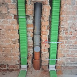 InstalHydroMax - Instalacja rur w ścianie z cegły, dwie zielone rury po bokach i szare rury w środku, mocowane metalowymi obejmami. Czerwona strzałka na ścianie.