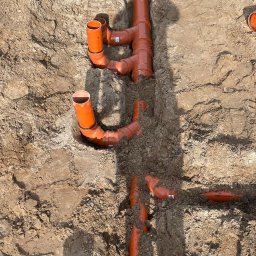 InstalHydroMax - Pomarańczowe rury kanalizacyjne ułożone w wykopie ziemnym, widok z góry. Elementy instalacji połączone kształtkami, widoczne fragmenty ziemi i narzędzi.