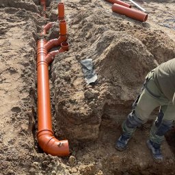 InstalHydroMax - Pomarańczowe rury kanalizacyjne ułożone w wykopie ziemnym, widoczne narzędzia i fragment sylwetki instalatora. Etap prac instalacyjnych na budowie.