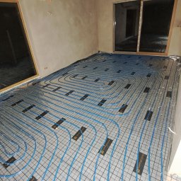 InstalHydroMax - Niebieskie rury ogrzewania podłogowego ułożone na srebrnej macie w surowym wnętrzu z oknami. Widok z góry na instalację przed wylaniem posadzki.