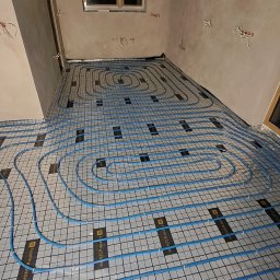 InstalHydroMax - Niebieskie rury ogrzewania podłogowego ułożone spiralnie na srebrnej macie w surowym wnętrzu z oknem i wystającymi kablami ze ścian.