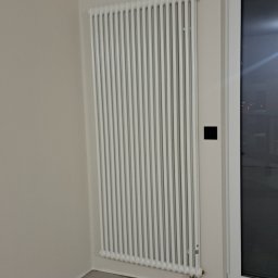 InstalHydroMax - Biały, żeliwny grzejnik żeberkowy zamontowany na ścianie w nowym, surowym wnętrzu. Widoczne przyłącza i fragment okna. Minimalistyczny design.