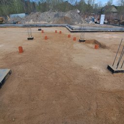 InstalHydroMax - Przygotowany teren pod budowę z fundamentami, widoczne wystające pręty zbrojeniowe i pomarańczowe rury. W tle hałda ziemi i drzewa.