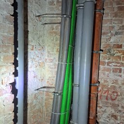 InstalHydroMax - Różne rury (szare, zielone, brązowe) zamocowane do ceglanej ściany w piwnicy w Warszawie. Widoczne uchwyty mocujące i izolacja termiczna na niektórych rurach.