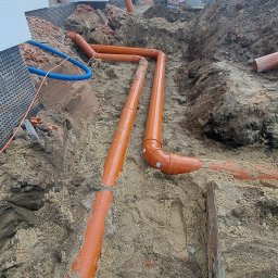 Oleh Matsiuk InstalHydroMax - Pomarańczowe rury kanalizacyjne ułożone w wykopie ziemnym przy budynku w trakcie budowy. Widoczne elementy izolacji fundamentów i niebieski wąż karbowany.