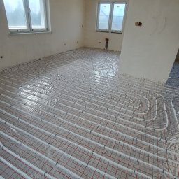 Oleh Matsiuk InstalHydroMax - Białe rury ogrzewania podłogowego ułożone na czerwonej siatce w surowym wnętrzu z dwoma oknami. Widok z góry na instalację przed wylaniem posadzki.