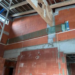 InstalHydroMax - Surowy stan budynku w trakcie budowy: ściany z czerwonej cegły, widoczne instalacje, drewniane belki stropowe i rusztowanie. Wnętrze w trakcie prac wykończeniowych.