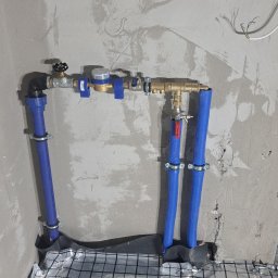 InstalHydroMax - Niebieskie rury instalacji wodnej z licznikiem, zamontowane na folii grzewczej podłogówki, na tle surowej ściany. Widoczne złącza i zawory.