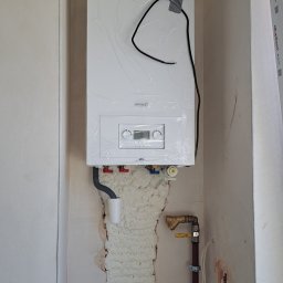 InstalHydroMax - Biały, nowy piec gazowy marki Brotje, jeszcze w folii ochronnej, zamontowany na ścianie. Widoczne rury, pianka montażowa i fragment okna. Ujęcie z bliska.
