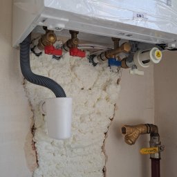 InstalHydroMax - Nowy piec gazowy, jeszcze w folii, z podłączonymi rurami i zaworami. Ściana wokół rur wypełniona pianką montażową. Kubek pod rurą odpływową.
