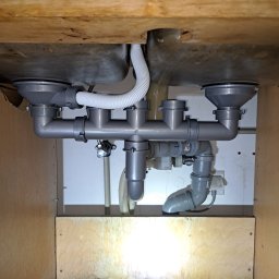 InstalHydroMax - Ukończona instalacja odpływowa pod podwójnym zlewem kuchennym, z szarych rur i białego węża, wewnątrz drewnianej szafki z widocznym gniazdkiem elektrycznym.