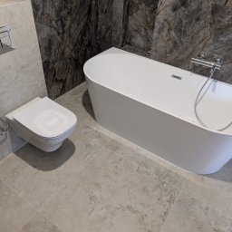 InstalHydroMax - Nowoczesna łazienka z białą wanną wolnostojącą i podwieszaną toaletą z bidetką, wykończona jasnymi płytkami na podłodze i ścianie oraz ciemnymi płytkami imitującymi marmur.