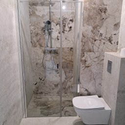InstalHydroMax - Nowoczesna łazienka z kabiną prysznicową z kwadratową deszczownicą i podwieszaną toaletą. Ściany zdobią jasne płytki imitujące marmur i szare, matowe kafle.