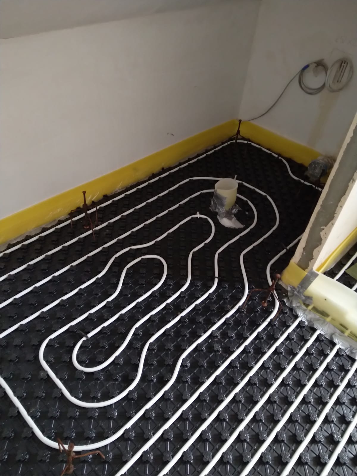 Białe rury ogrzewania podłogowego ułożone na czarnej macie z żółtą folią na ścianach, widoczne elementy montażowe i plastikowy kubek.