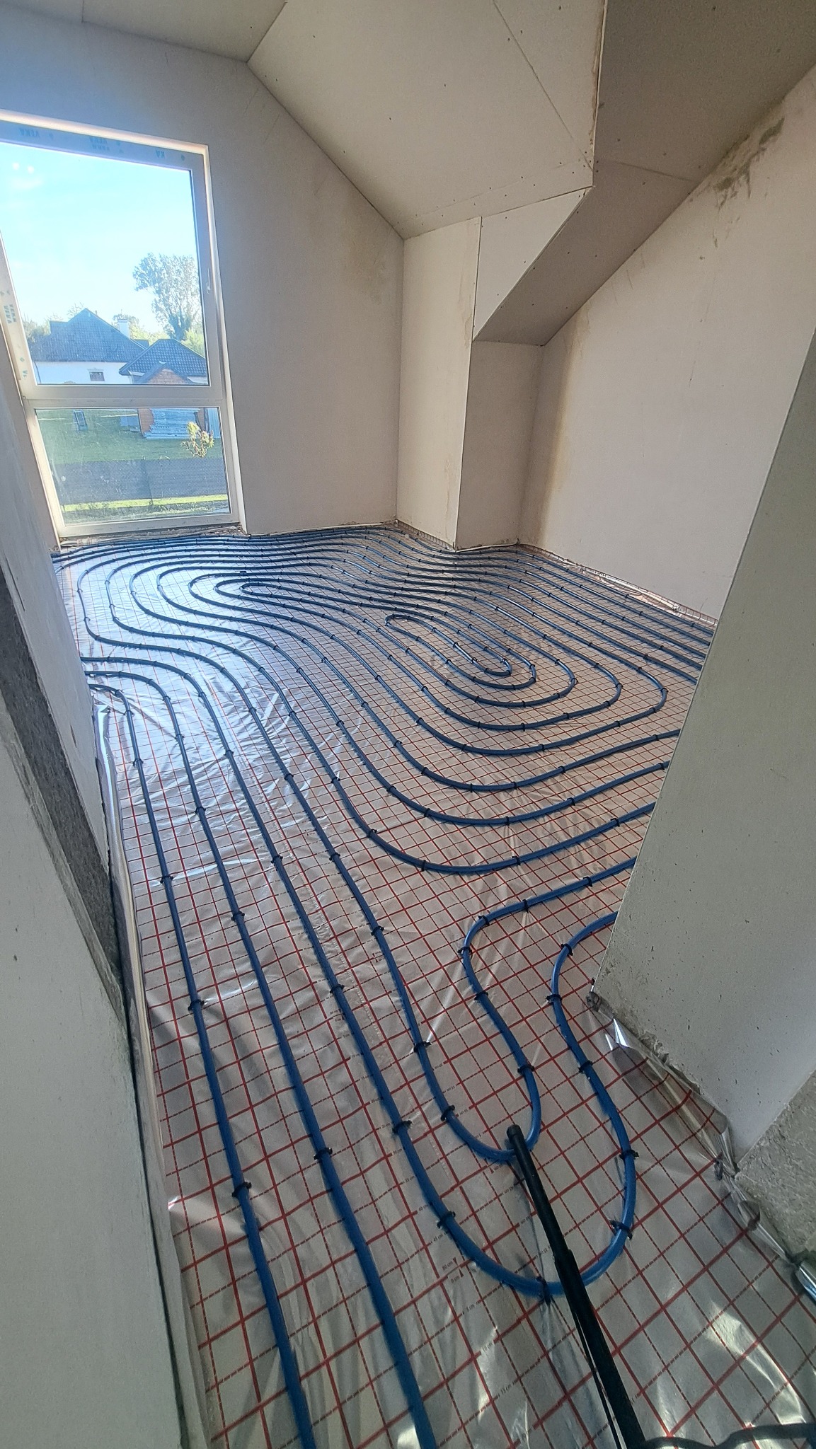 Układanie niebieskich rur ogrzewania podłogowego na folii z siatką w surowym wnętrzu poddasza z oknem. Widok z góry, częściowo widoczny czarny element instalacji.