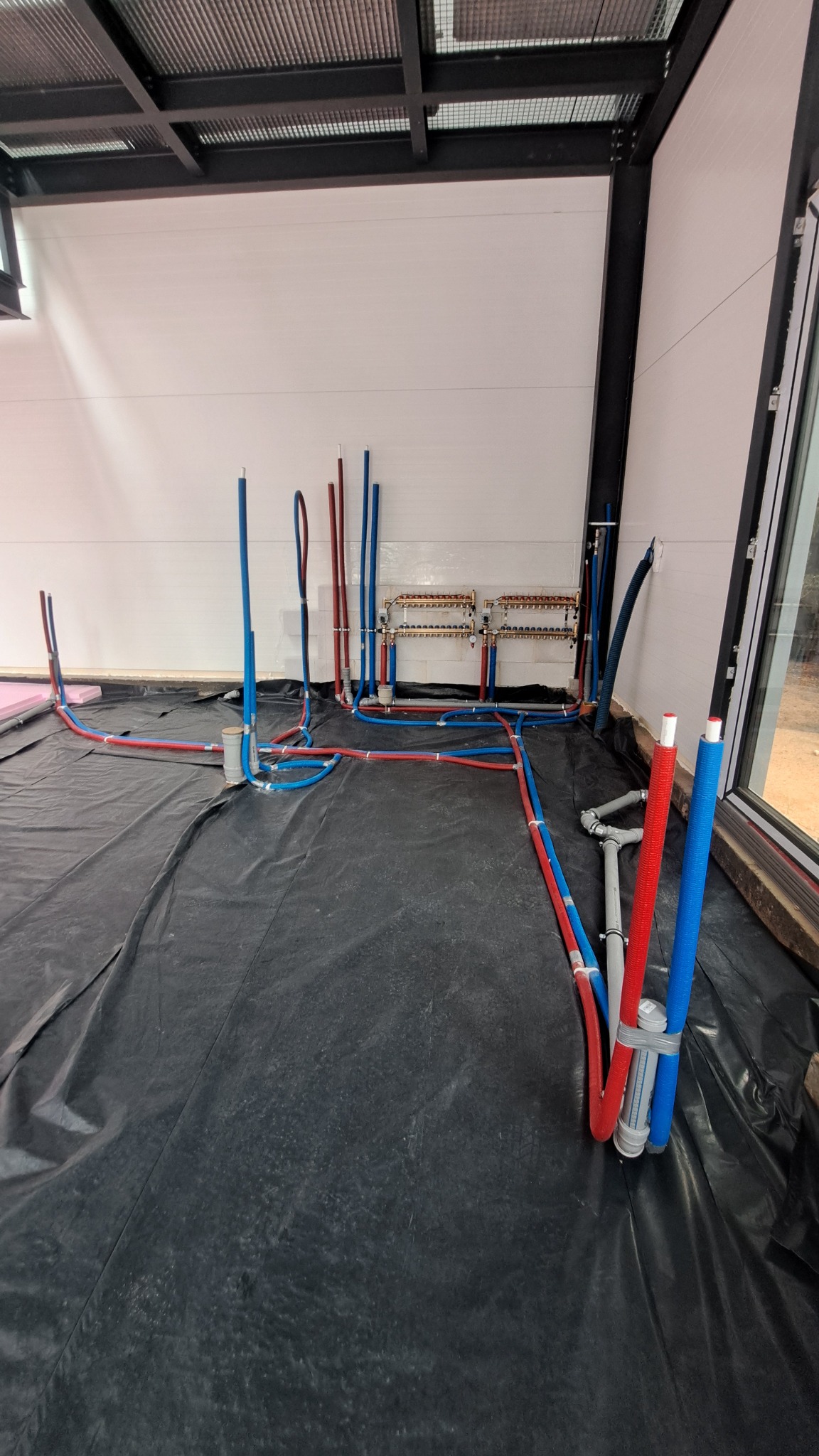 Instalacja ogrzewania podłogowego w nowym budynku. Widoczne rury w kolorach niebieskim i czerwonym, rozłożone na czarnej folii izolacyjnej, z rozdzielaczem na ścianie.