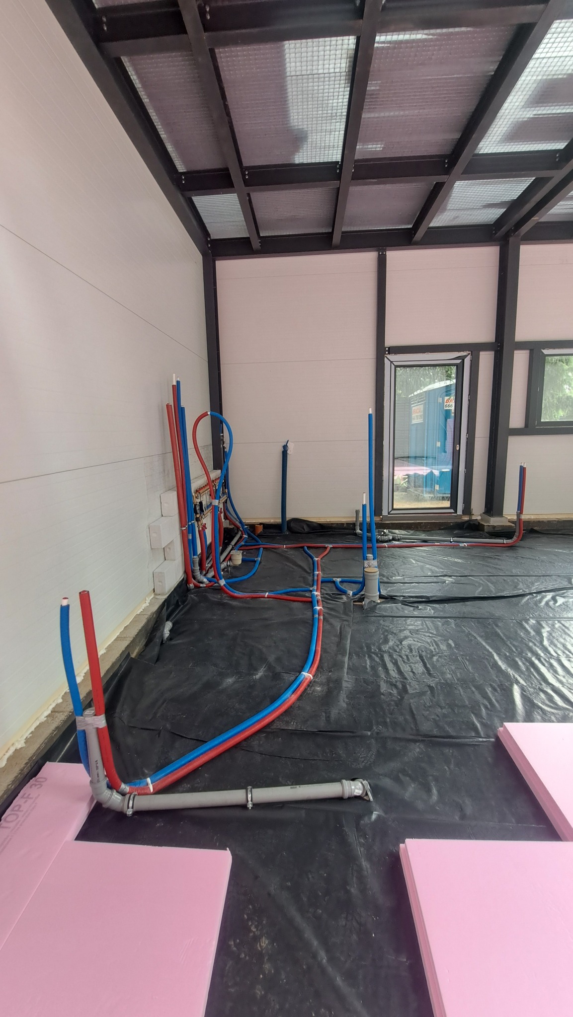 Instalacja rur grzewczych (czerwone i niebieskie) na czarnej folii, z widocznymi fragmentami różowych płyt izolacyjnych i elementami konstrukcji budynku.