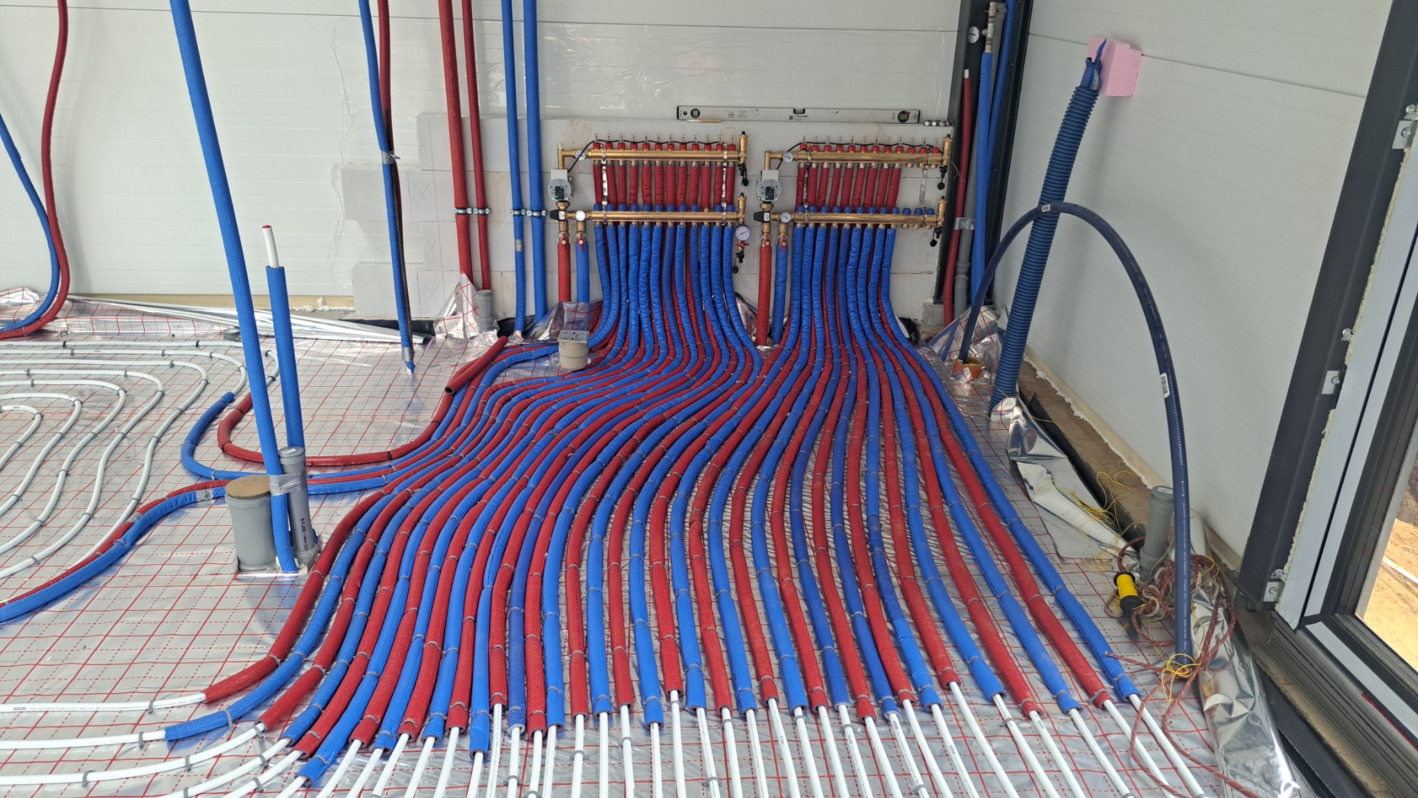 Instalacja ogrzewania podłogowego z rozdzielaczem, rury w izolacji czerwono-niebieskiej, białe rurki grzewcze na folii z rastrem, widok z góry, w tle białe ściany.