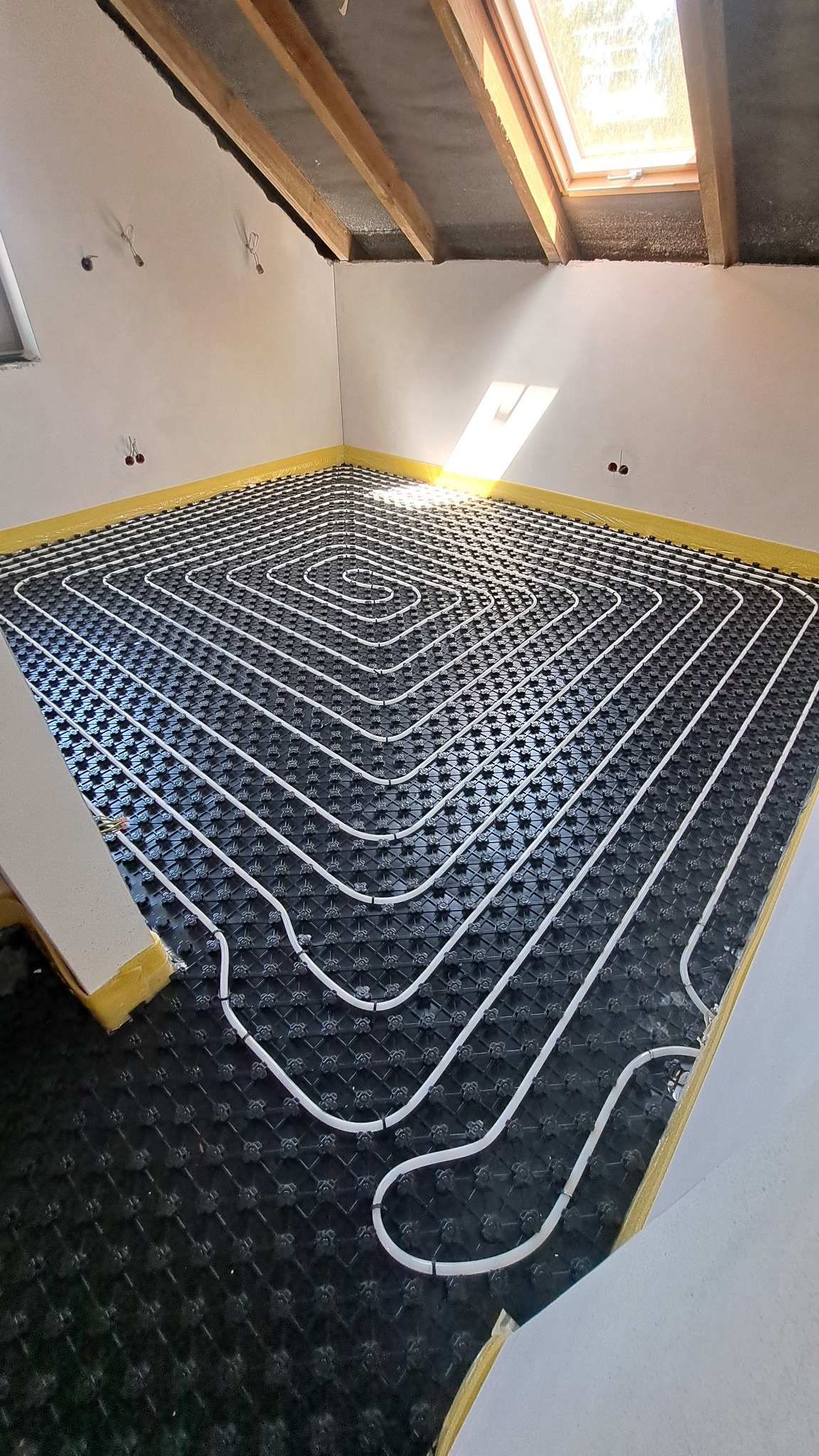 Białe rurki ogrzewania podłogowego ułożone spiralnie na czarnej macie w pokoju z oknem dachowym. Żółta taśma na styku ściany i podłogi.