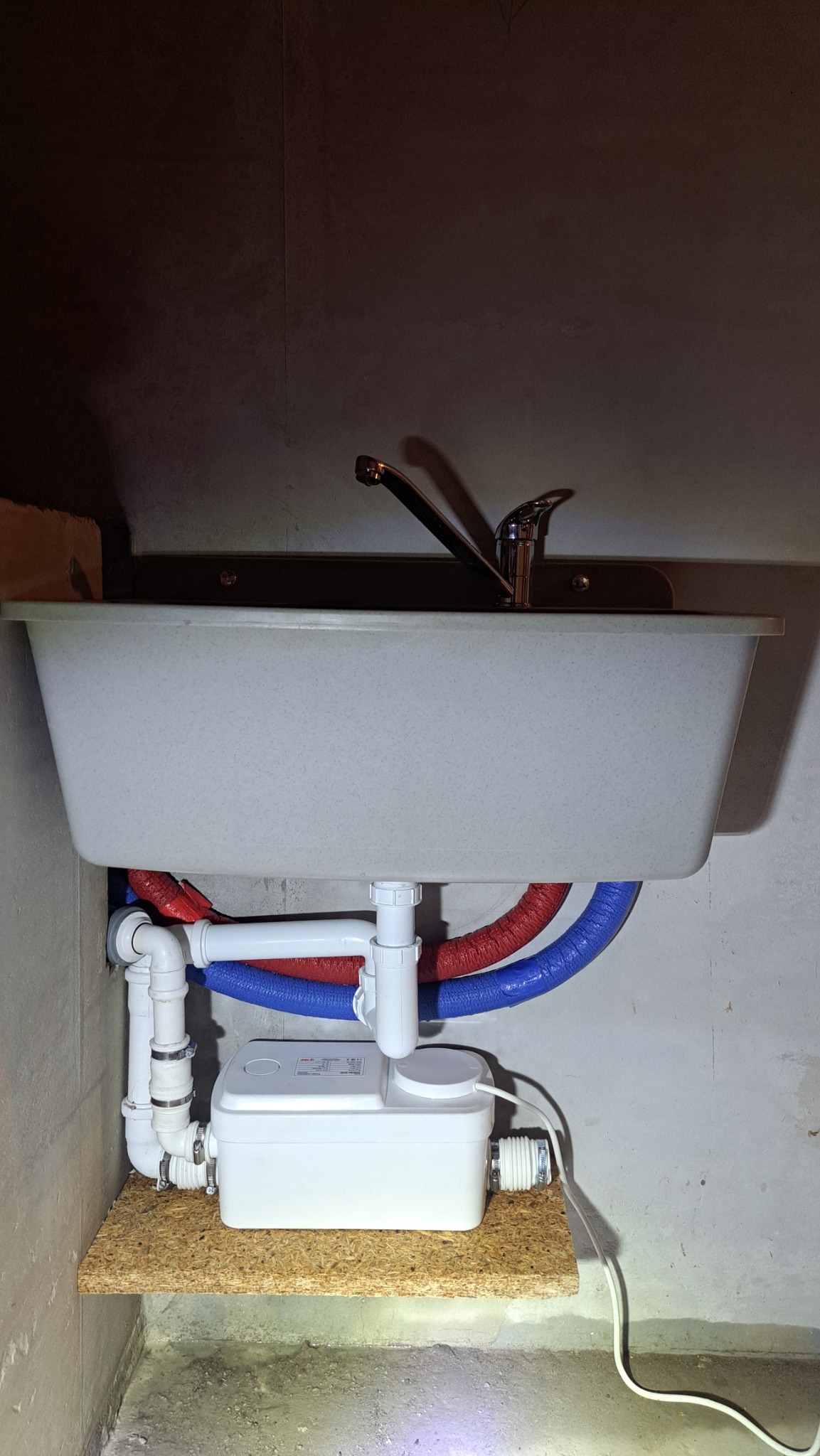 Szara umywalka z baterią i podłączonymi rurami (czerwona i niebieska) oraz białą pompą sanitarną na drewnianej półce w surowym wnętrzu.