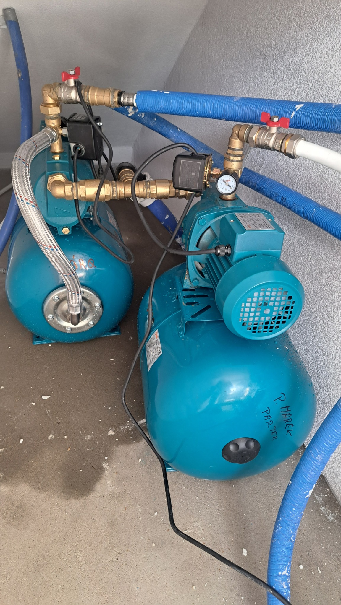 Dwa turkusowe hydrofory z manometrem i rurami miedzianymi oraz niebieskimi w izolacji, umieszczone w pomieszczeniu z szarą ścianą i betonową podłogą.