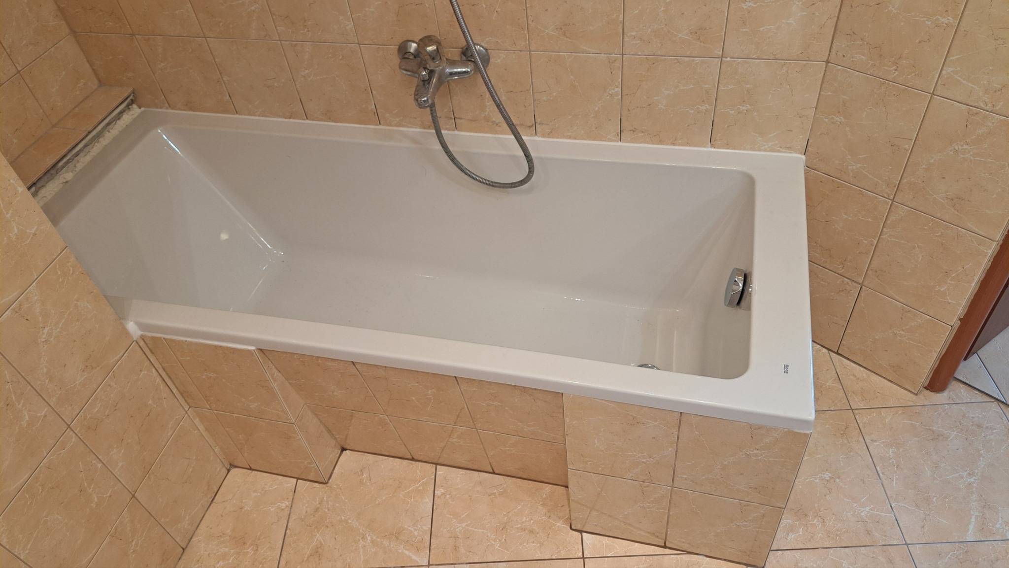 Biała wanna obudowana beżowymi płytkami w łazience. Widoczna bateria i słuchawka prysznicowa. Ujęcie z góry, ukazujące estetyczne wykończenie.