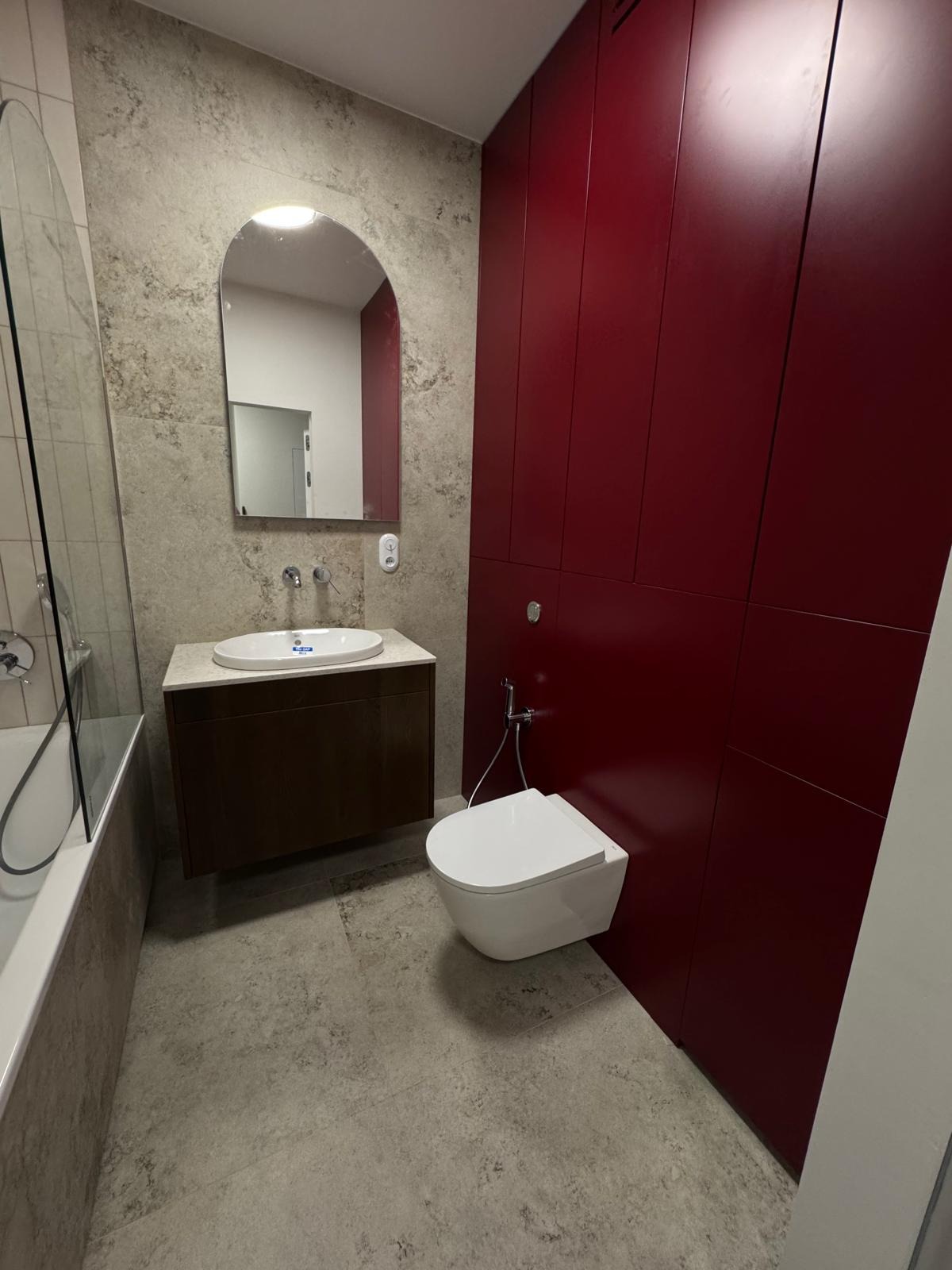 Nowoczesna łazienka z umywalką na szafce, podwieszaną toaletą z bidetką i dużymi, czerwonymi szafami w zabudowie. Lustro owalne z podświetleniem, płytki imitujące kamień.