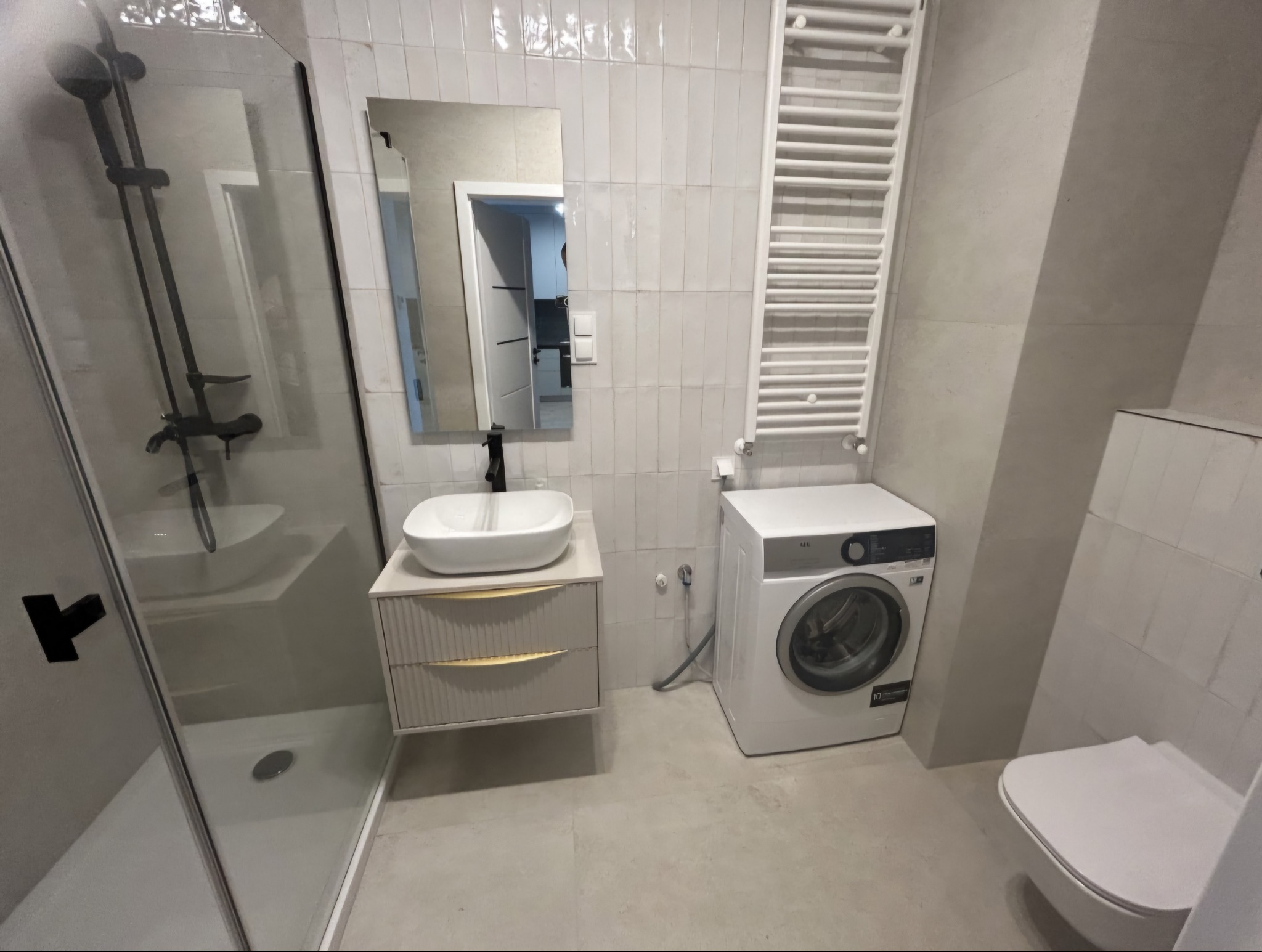 Biała łazienka z kabiną prysznicową, umywalką na szafce z frezowanymi frontami, grzejnikiem drabinkowym i pralką. Nowoczesne wykończenie.