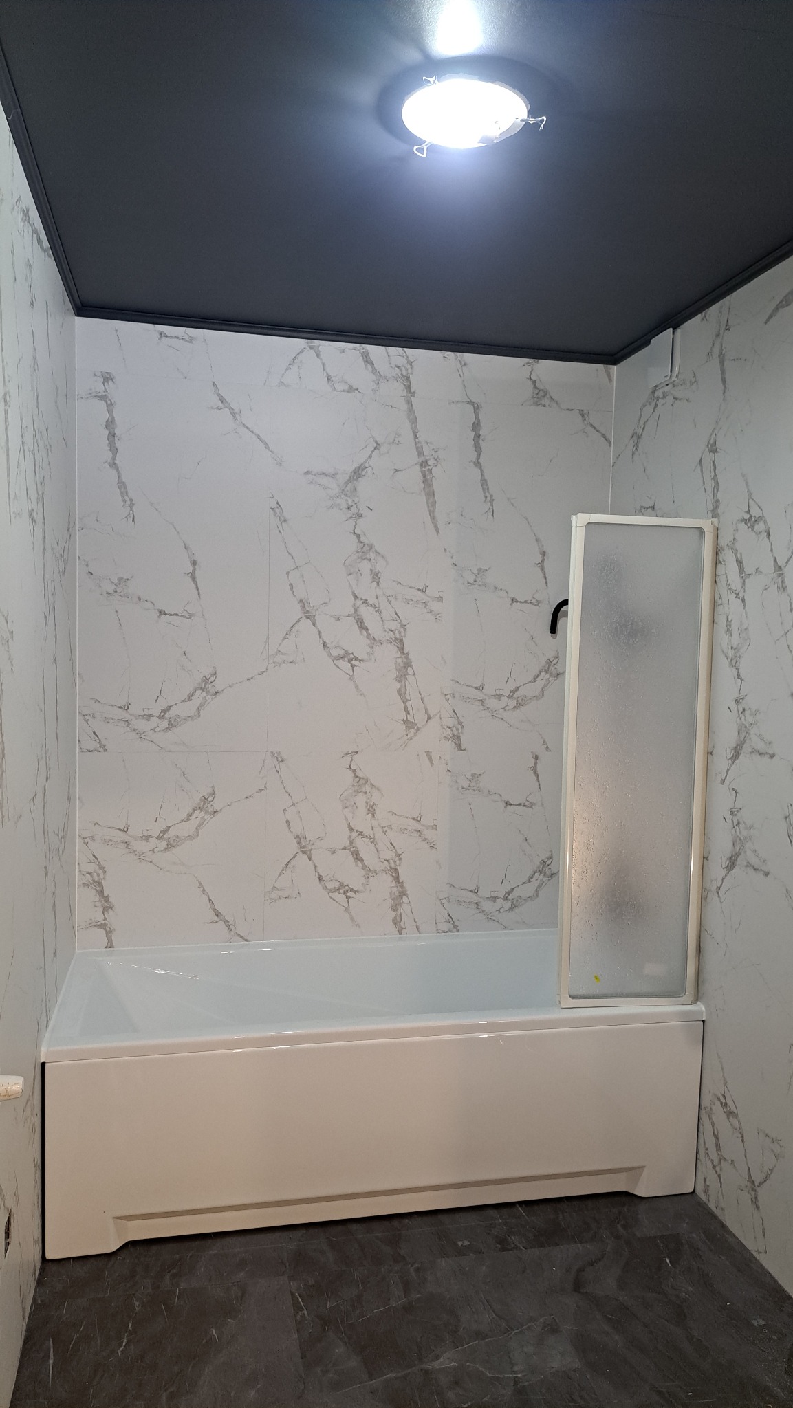 Biała wanna z parawanem nawannowym na tle marmurowych płytek i ciemnego sufitu. Widoczne oświetlenie sufitowe. Nowoczesna aranżacja łazienki po remoncie.