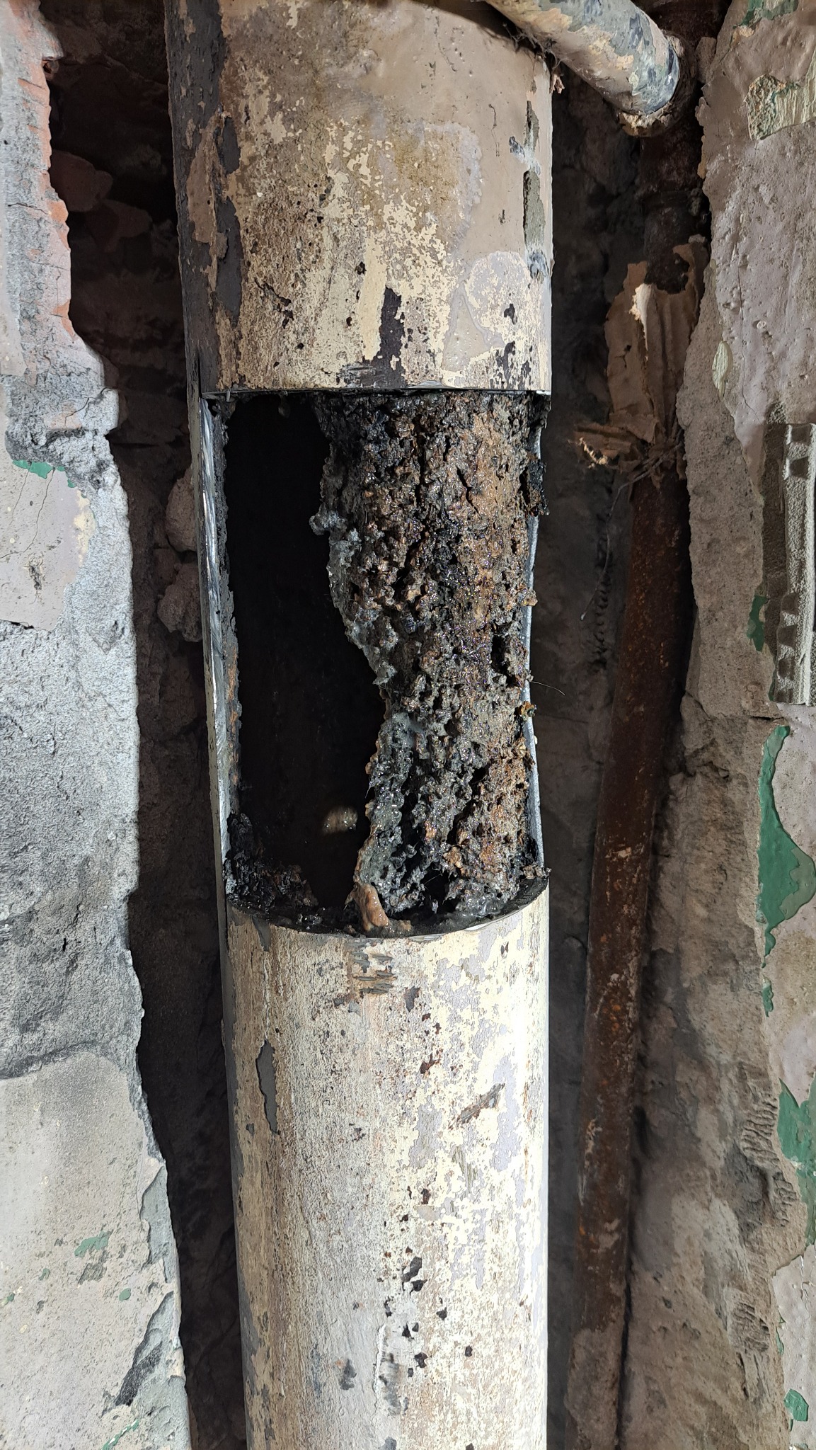 Zniszczona, skorodowana rura kanalizacyjna z widocznym nagromadzeniem osadów wewnątrz, umieszczona w ścianie z odpadającym tynkiem. Widoczne ślady korozji i uszkodzeń.