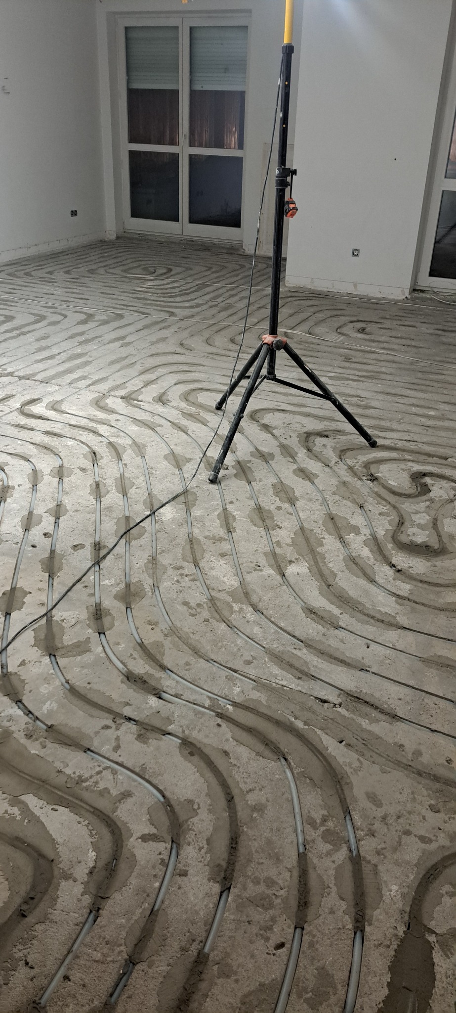 Szare rury ogrzewania podłogowego ułożone w betonowej wylewce, widoczne w surowym wnętrzu z oknami i lampą budowlaną na trójnogu. Etap prac przed zalaniem posadzki.