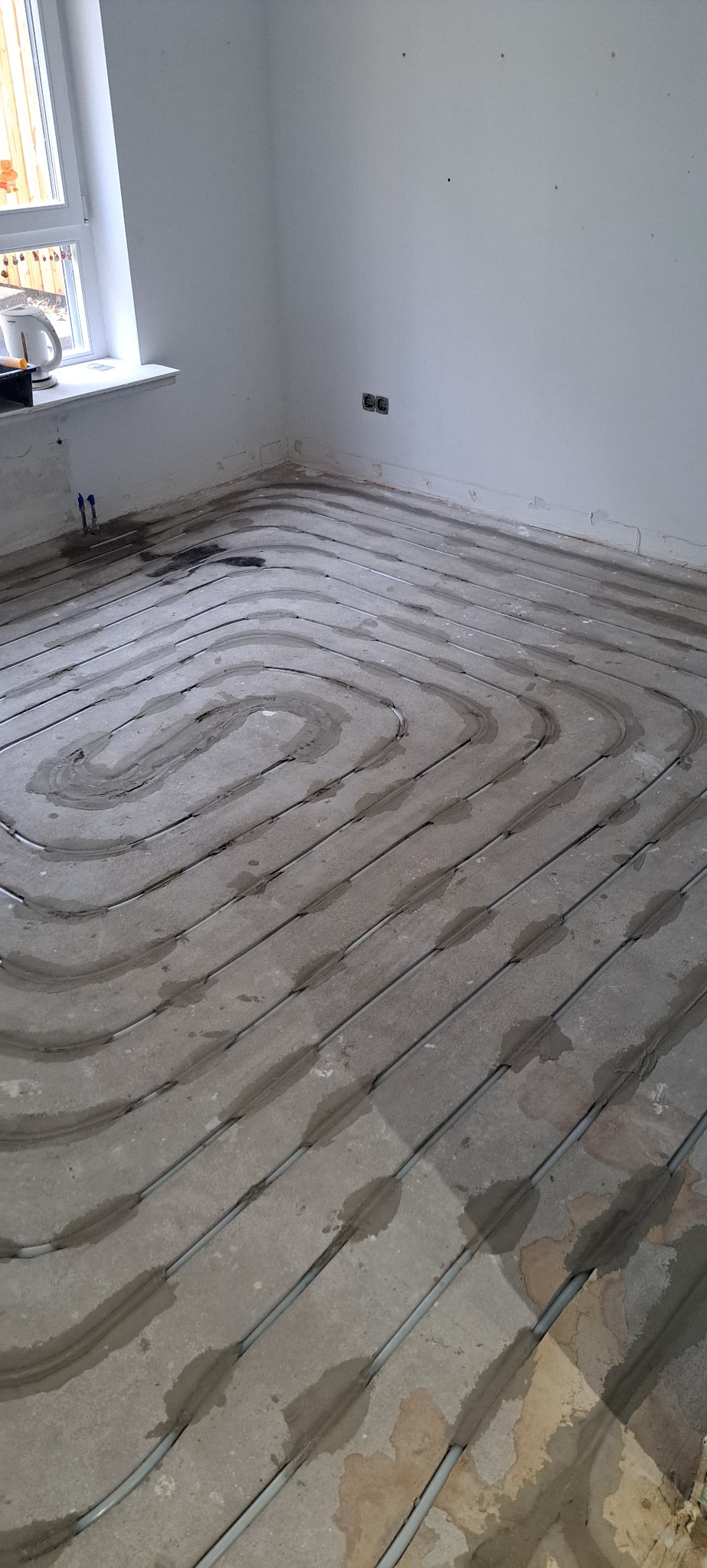 Szare rury ogrzewania podłogowego ułożone spiralnie na betonowej wylewce w jasnym pomieszczeniu z oknem. Widoczne ślady zaprawy i niedoskonałości podłoża.