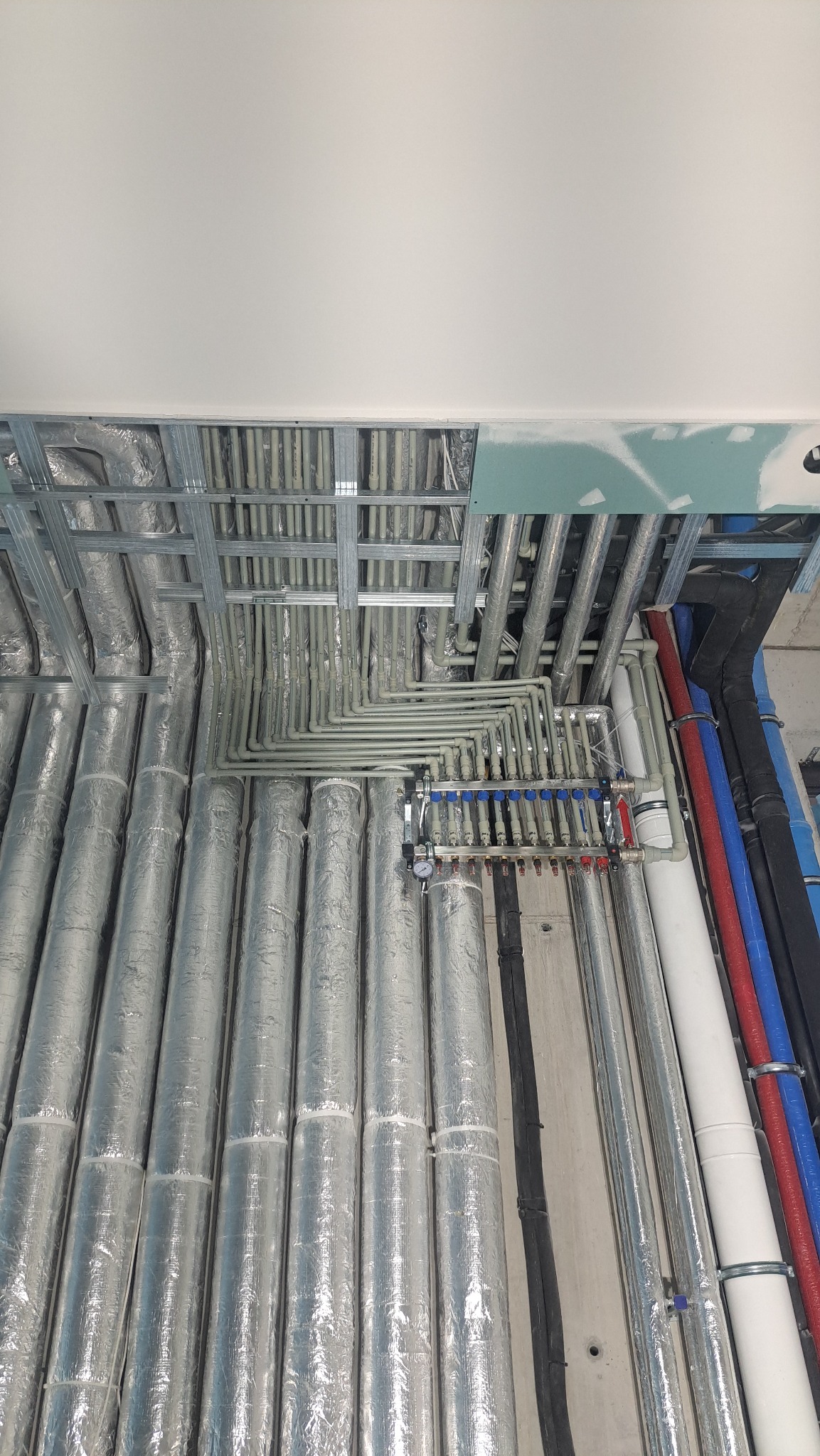 Rozbudowana instalacja grzewcza pod stropem z licznymi rurami w izolacji, rozdzielaczem i elementami montażowymi. Widoczne rury w różnych kolorach.