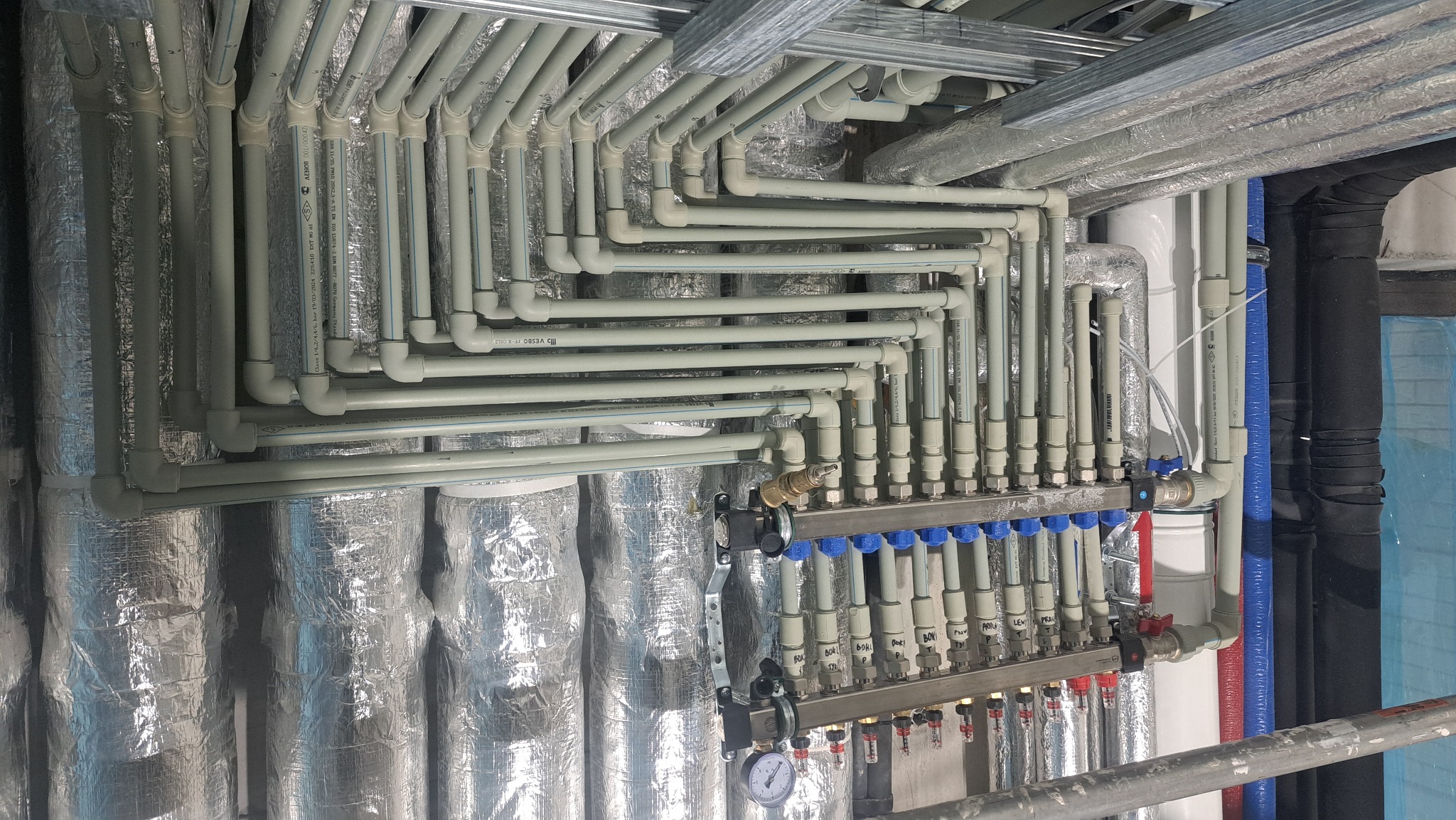 Rozbudowana instalacja ogrzewania podłogowego z licznymi rurami i rozdzielaczem, widoczna pod sufitem, z izolacją termiczną na ścianach i elementami konstrukcyjnymi.