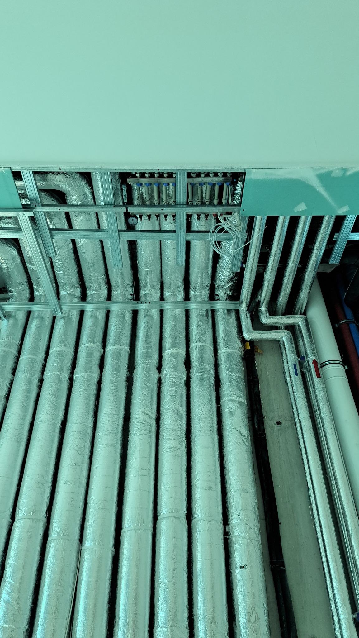 Równolegle ułożone, zaizolowane rury instalacji pod sufitem w trakcie montażu, widoczne elementy konstrukcji sufitu podwieszanego i rozdzielacz z zaworami.