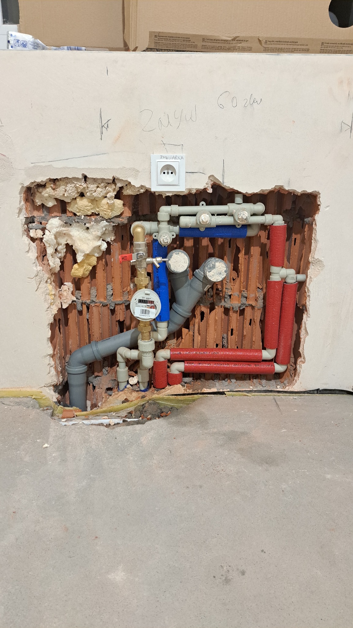 Odsłonięta instalacja wodna w ścianie z widocznymi rurami szarymi i czerwonymi, zaworami, licznikiem wody i gniazdkiem elektrycznym powyżej. Widoczne ślady po kuciu.