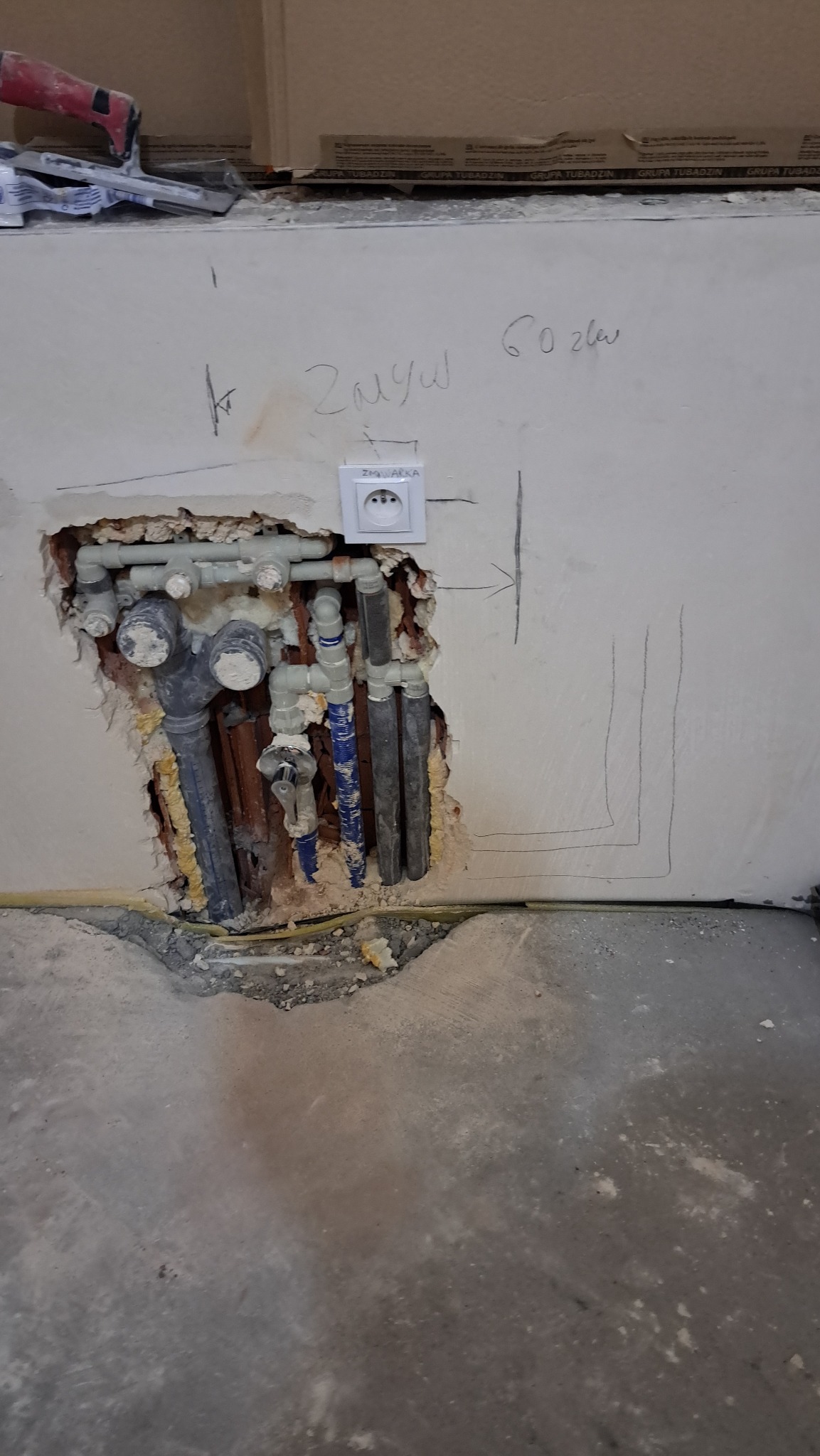 Odsłonięte rury instalacji wodnej i elektryczne gniazdko w ścianie podczas remontu. Widoczne oznaczenia ołówkiem na ścianie i pianka montażowa.