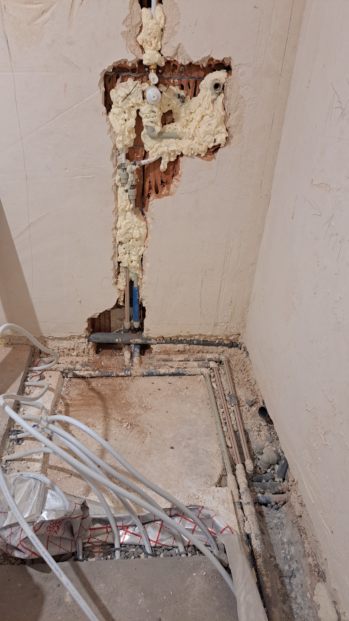 Ściana z wykuciem i pianką montażową wokół rur, instalacja podłogowa w trakcie montażu. Widoczne rury i folia izolacyjna na podłodze. Prace hydrauliczne w surowym wnętrzu.