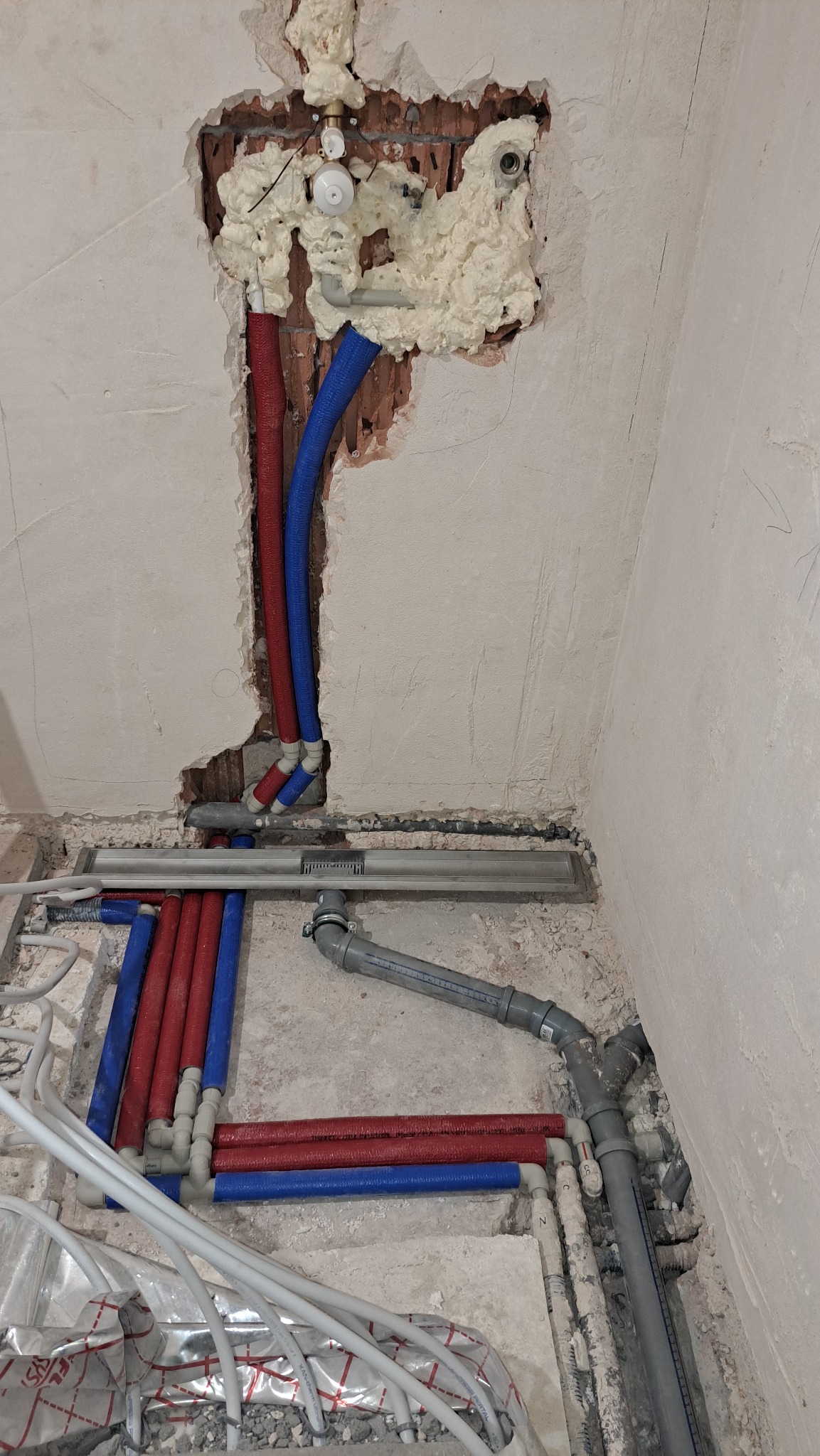 Instalacja wodna w trakcie montażu: czerwone i niebieskie rury prowadzone w ścianie i podłodze, zabezpieczone pianką montażową, widoczny odpływ liniowy.