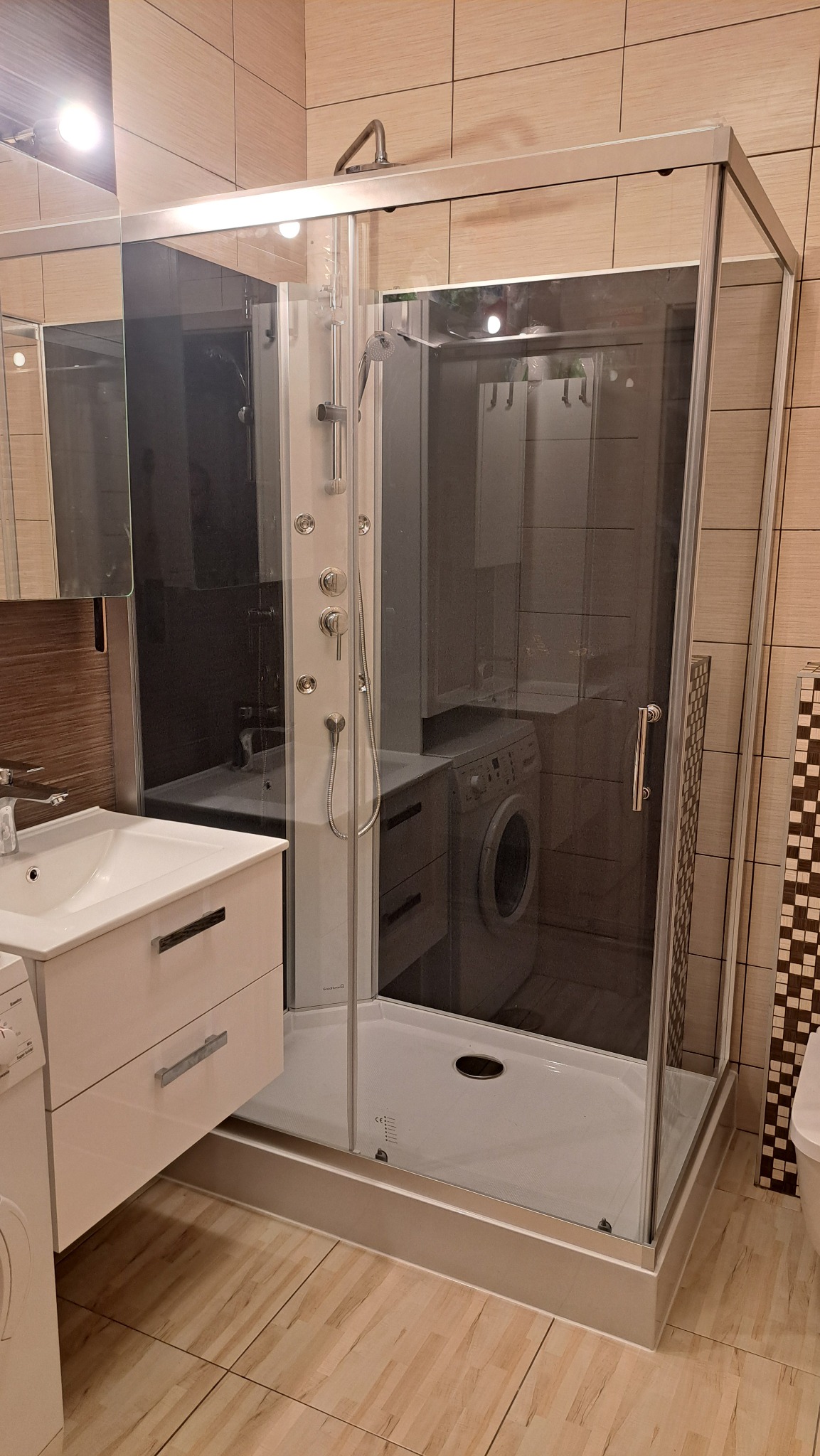 Nowoczesna kabina prysznicowa z hydromasażem, umywalką i pralką w tle, w łazience wykończonej płytkami w odcieniach beżu i brązu. Widok z perspektywy podłogi.