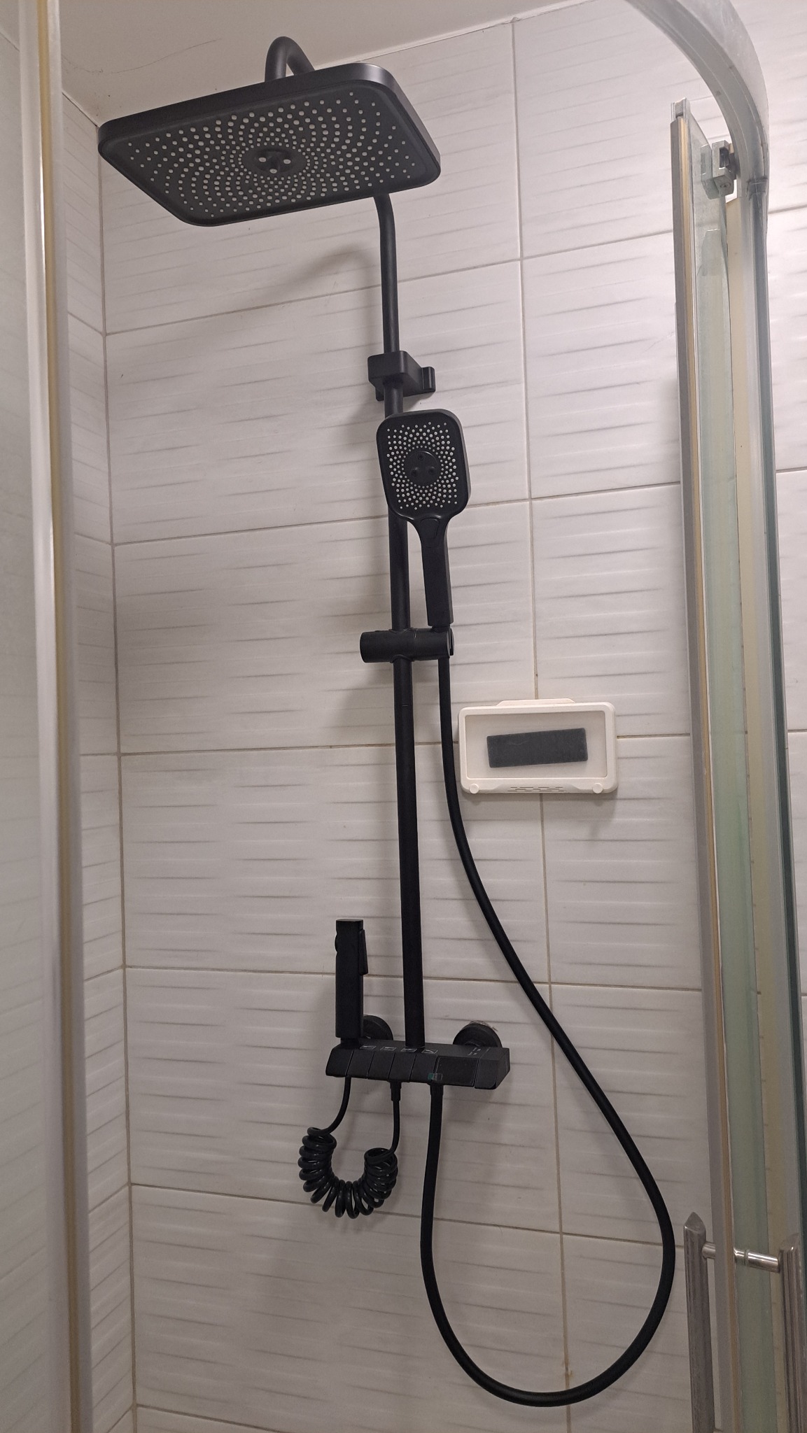 Czarny, kwadratowy zestaw natryskowy z deszczownicą i słuchawką, zamontowany na białej, wzorzystej ścianie kabiny prysznicowej. Widoczny dozownik na mydło.