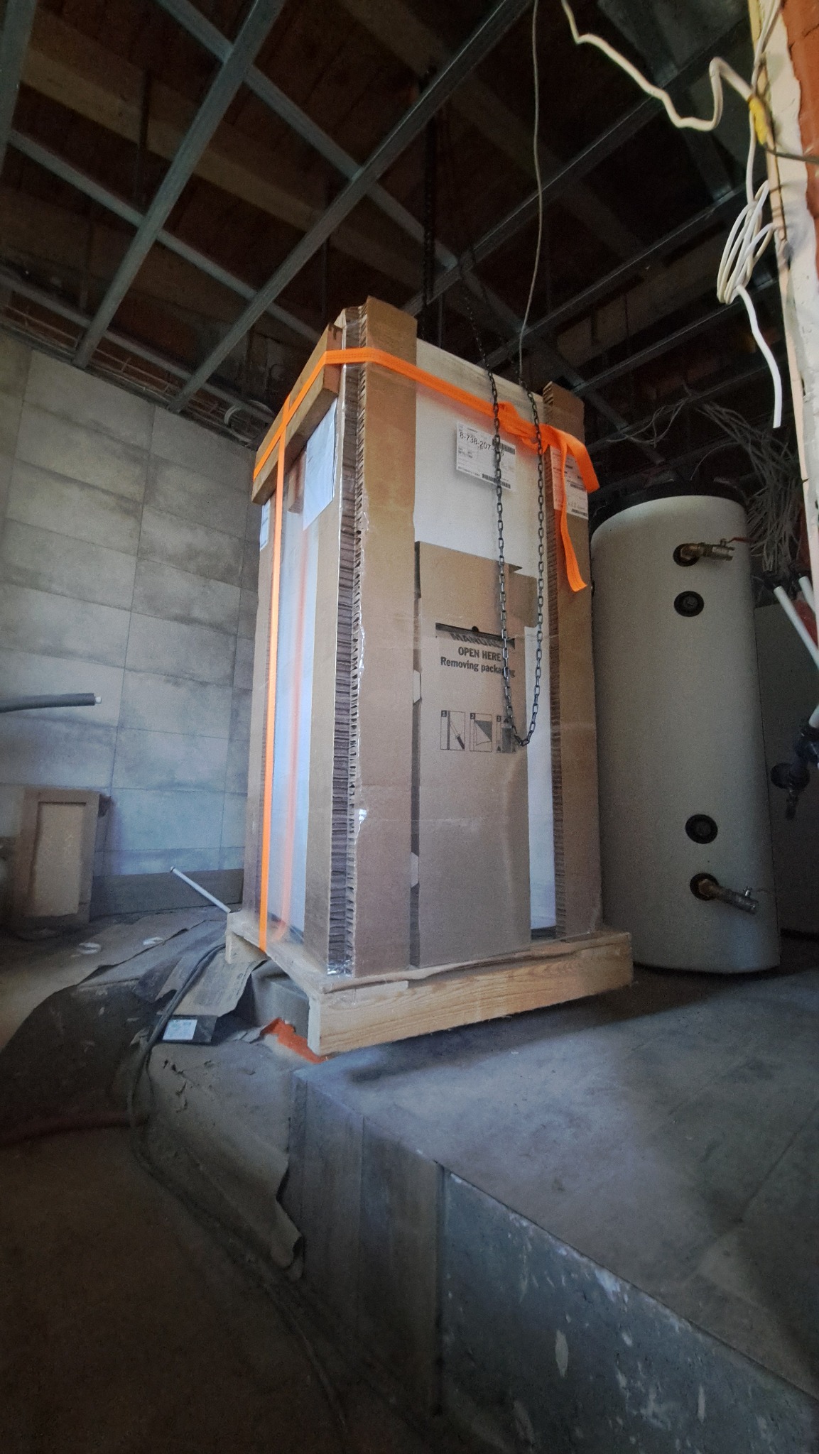 Nowy, zapakowany bojler na palecie, zabezpieczony pasami transportowymi, podwieszony na łańcuchu w surowym wnętrzu z widoczną instalacją.