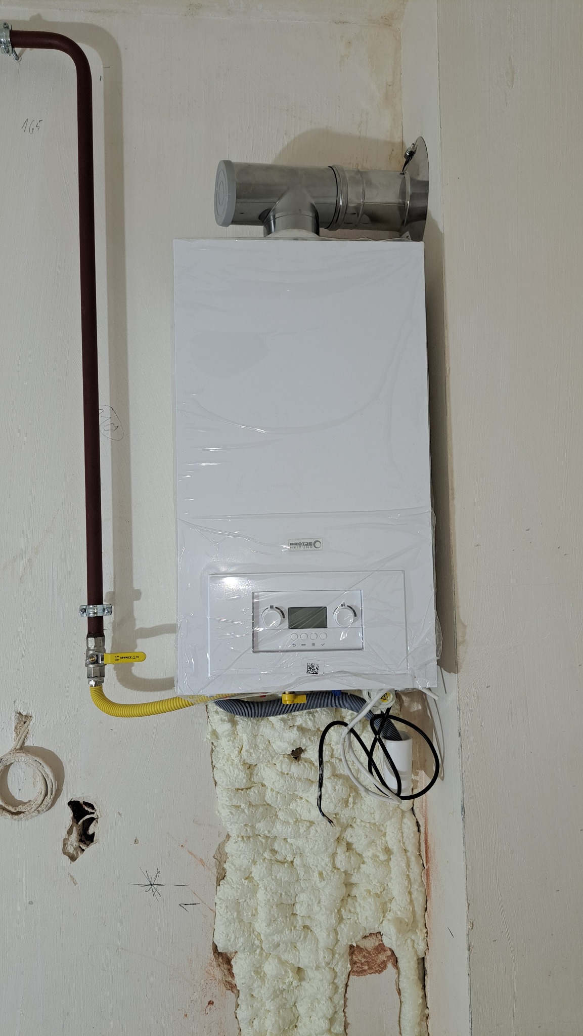 Biały, nowy piec gazowy marki Brötje, jeszcze w folii ochronnej, zamontowany na ścianie z widoczną pianką montażową i rurami. Widoczny fragment instalacji gazowej.
