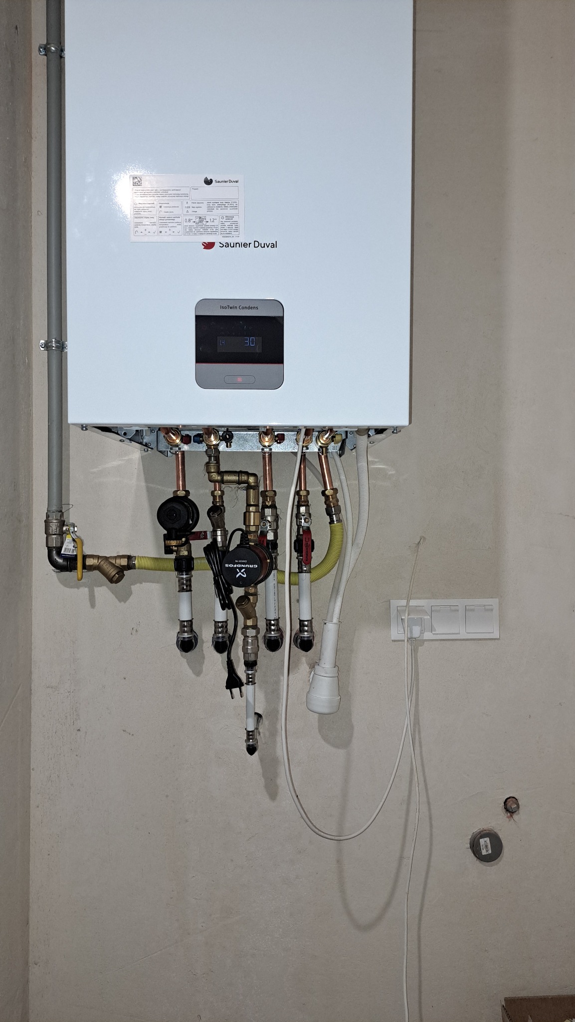 Nowoczesny, wiszący piec gazowy Saunier Duval z podłączeniami hydraulicznymi i elektrycznymi na surowej, betonowej ścianie. Widoczne rury miedziane i elementy instalacji.