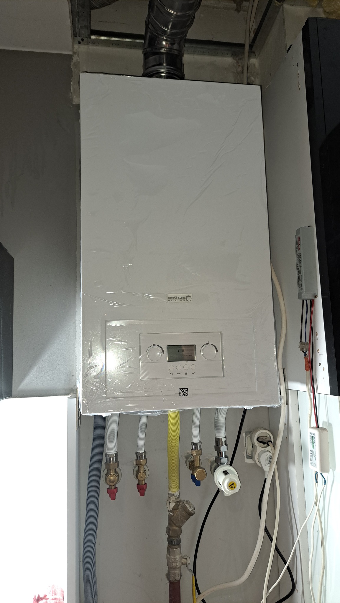 Nowy, biały piec gazowy marki Brötje, jeszcze w folii ochronnej, z podłączonymi rurami i zaworami, zamontowany w pomieszczeniu technicznym. Widoczne przewody elektryczne.